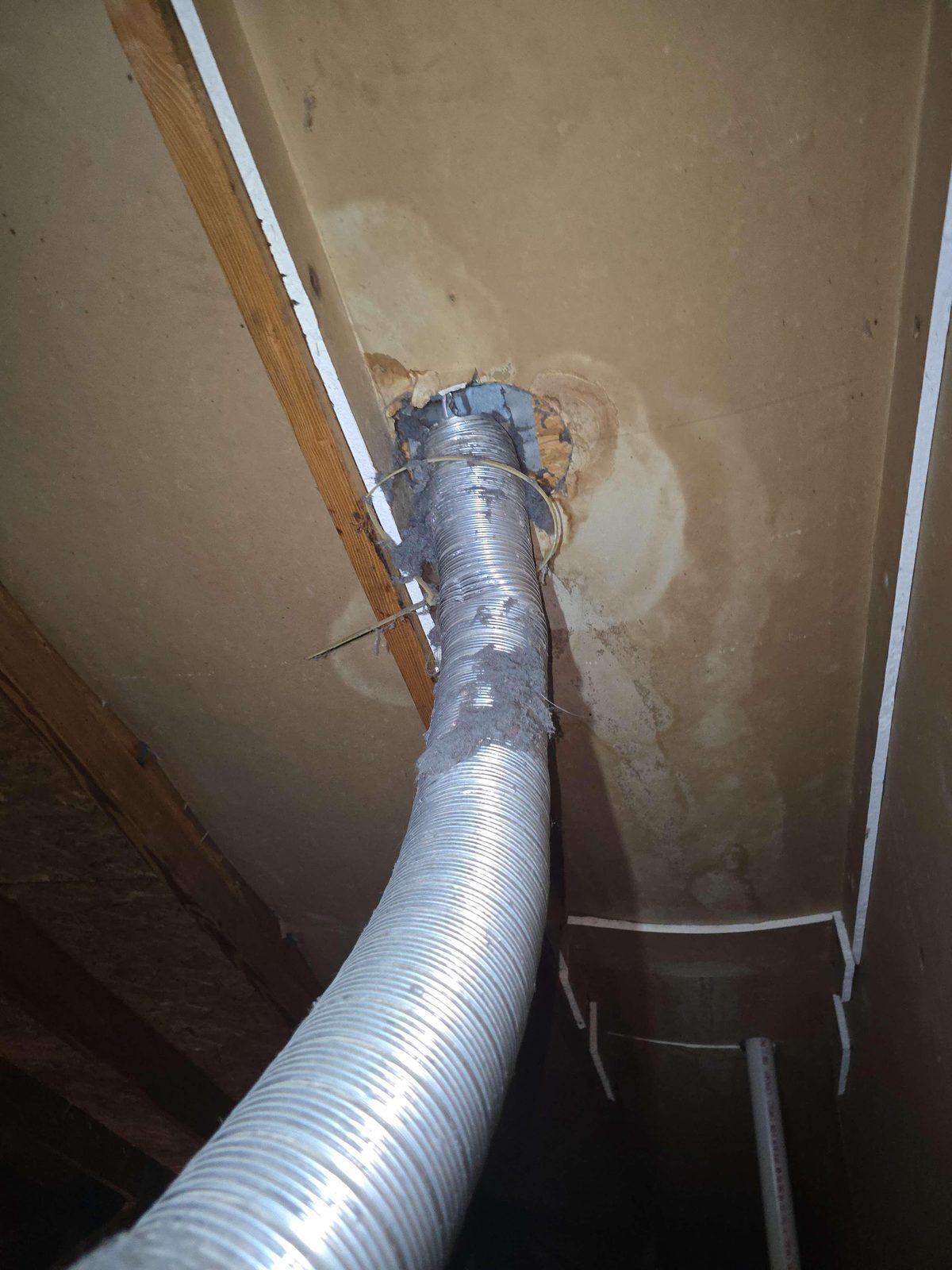 Attic space showing a dryer exhaust duct with an open, unconnected end and a visible accumulation of lint on nearby attic surfaces and insulation.