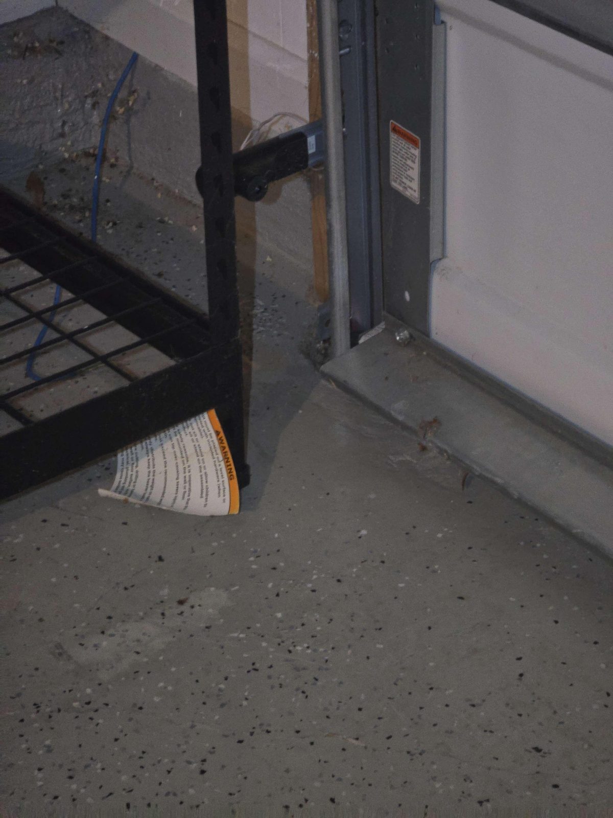 A garage door photo-eye sensor mounted on a metal bracket along the inside of a garage door frame, positioned several inches above the concrete floor.