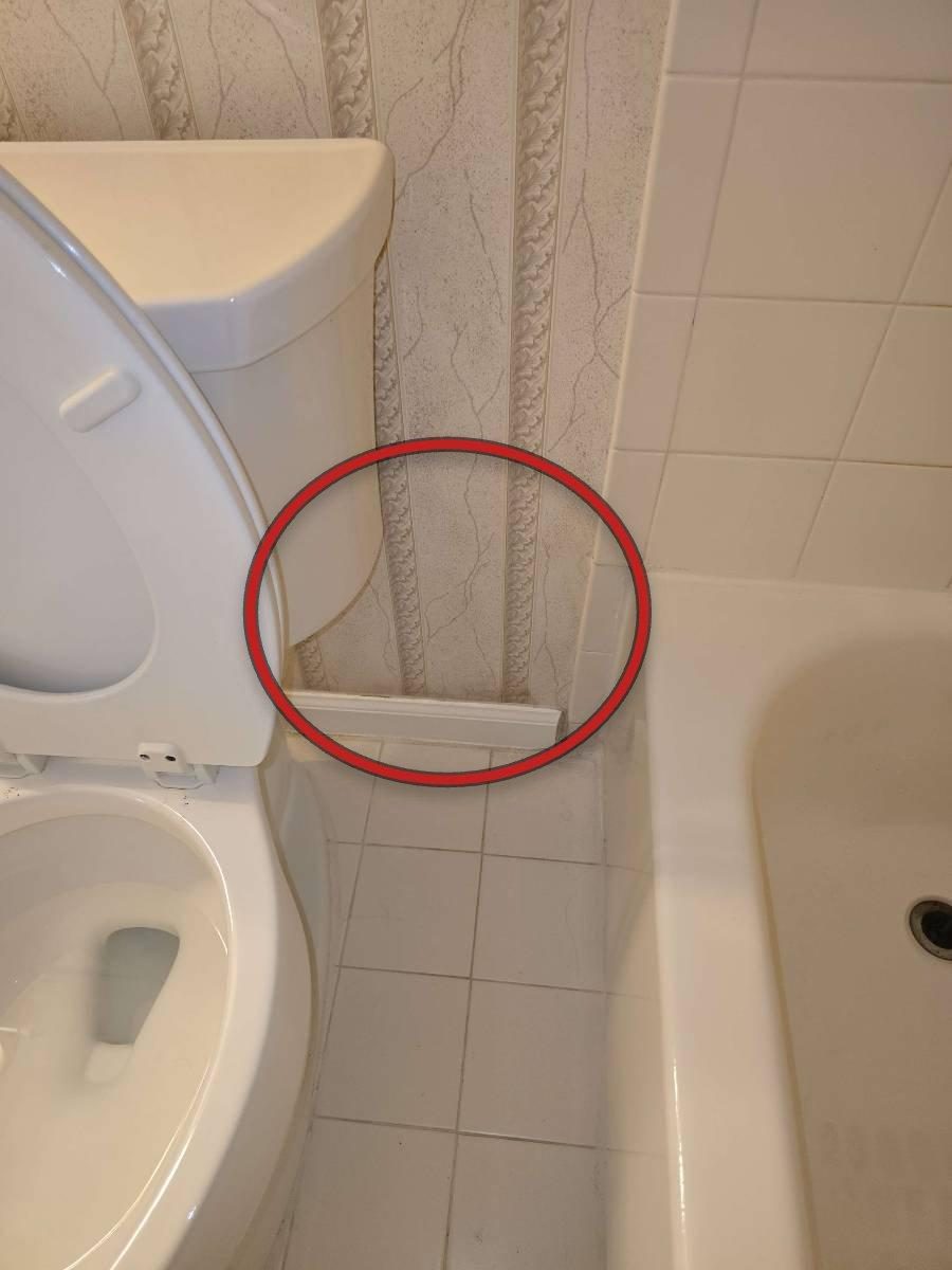A moisture meter pressed against white drywall beside a bathtub surround, with the digital display showing a high reading