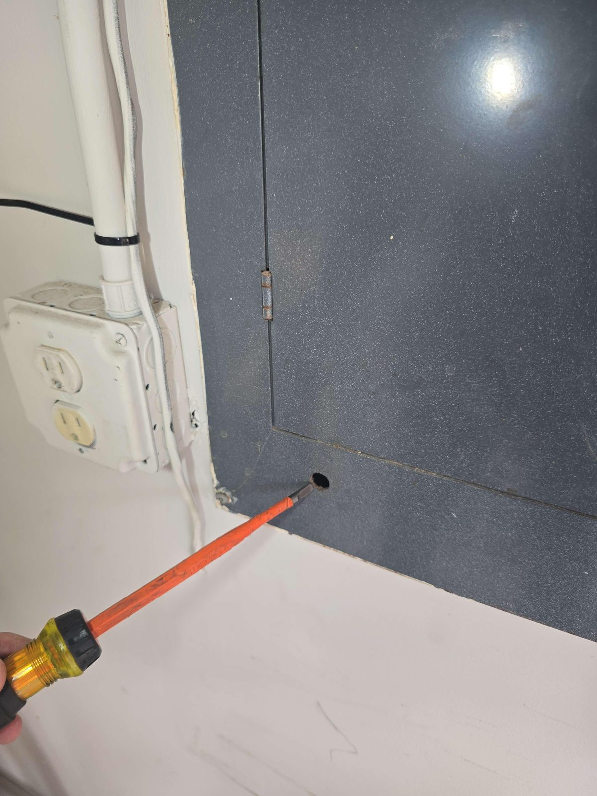A residential electrical panel dead-front cover — the metal plate that sits over the breakers — with a hole drilled through it.