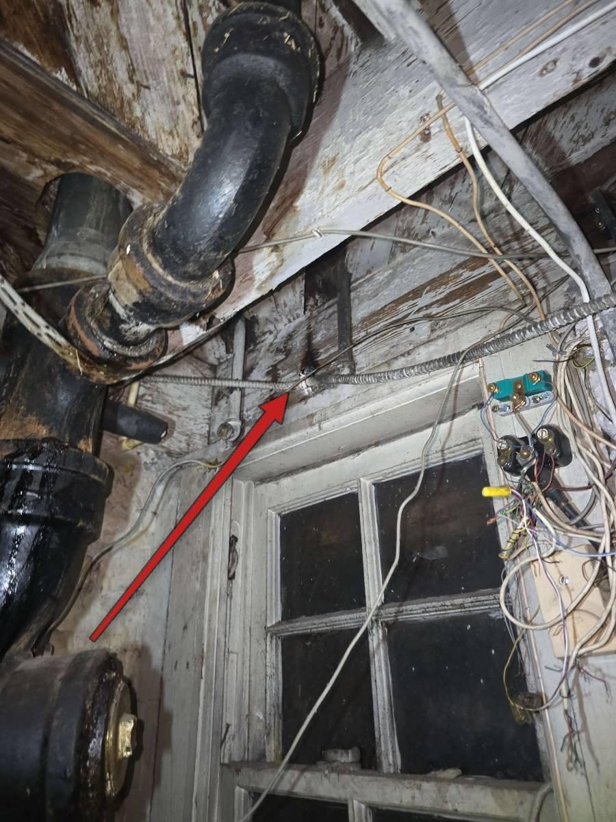 Close-up of a flexible metal conduit where it meets a wood framing member in a basement; the conduit strap or fastener is burned away and the surrounding wood is visibly charred black