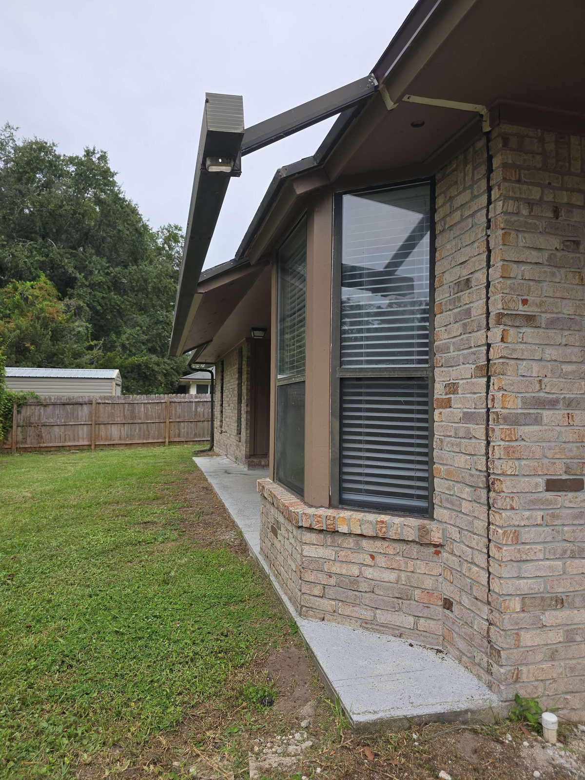 Partially damaged aluminum screen enclosure framing at the rear of a home, showing gaps where sections are missing, with a super gutter visible above lacking a downspout at its end.